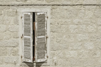 A weathered wooden window shutter is slightly open against a textured stone wall. The wall is made of large, roughly-cut gray stones, and the shutter is painted white with visible signs of age and discoloration.