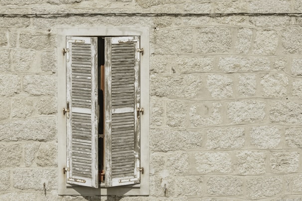 A weathered wooden window shutter is slightly open against a textured stone wall. The wall is made of large, roughly-cut gray stones, and the shutter is painted white with visible signs of age and discoloration.