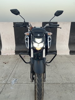 A motorcycle is parked on a concrete surface, positioned in front of a black-and-white striped barrier. The motorcycle is equipped with a headlight that is turned on, and the front view of the bike is visible, including its handlebars and mirrors.