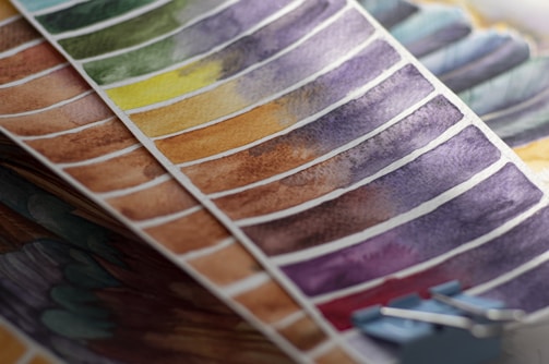 Brightly colored watercolor paint swatches arranged in rows on paper. The swatches vary in hue from green and yellow to brown and purple, showing a range of gradients. A blue binder clip is partially visible at the edge, holding some of the sheets together.