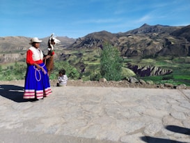 A woman in traditional clothing stands beside a llama on a paved path overlooking a scenic mountainous landscape with terraced fields. A child sits nearby, facing the view.