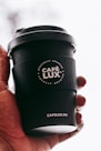 A hand is holding a black coffee cup with the logo 'CAFÉ LUX*' prominently displayed. The cup also features the text 'Ethically Sourced, Locally Roasted' and the website 'CAFELUX.MU'. The background is softly blurred, emphasizing the cup.