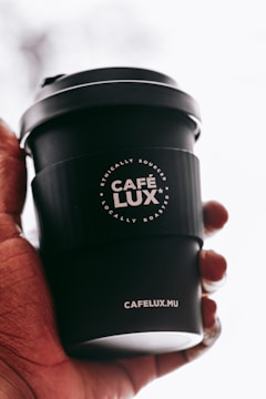 A hand is holding a black coffee cup with the logo 'CAFÉ LUX*' prominently displayed. The cup also features the text 'Ethically Sourced, Locally Roasted' and the website 'CAFELUX.MU'. The background is softly blurred, emphasizing the cup.