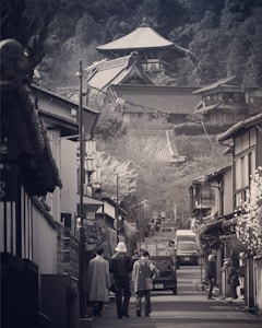 A narrow street is flanked by traditional Japanese buildings with sloping roofs. People are walking along the street, and there are a few cars parked and driving. In the background, several traditional Japanese temples with intricate roofs can be seen, nestled in a hilly, forested area.