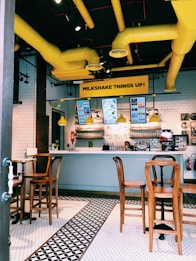 A modern caf&eacute; interior features a sleek white-tiled counter with a mix of vintage and modern elements. Bright yellow ducts contrast with the ceiling, and wooden high chairs are neatly arranged around small tables. The menu is displayed on screens above the counter, where a person is seen standing. The floor features a classic black and white geometric tile pattern.