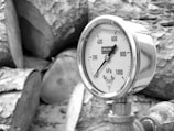 A close-up view of a pressure gauge mounted on a pipe. The gauge displays measurements in kilopascals, ranging from 0 to 1000 kPa. The reading is around 0. The background consists of large, cut logs with a textured surface.