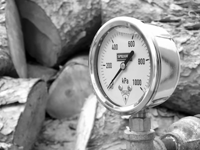 A close-up view of a pressure gauge mounted on a pipe. The gauge displays measurements in kilopascals, ranging from 0 to 1000 kPa. The reading is around 0. The background consists of large, cut logs with a textured surface.