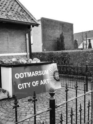 A black and white image of an outdoor scene featuring a sign that reads 'Ootmarsum City of Art' leaning against a building with brick walls and a tiled roof. The scene includes a paved area with a decorative iron fence in the foreground and neatly trimmed hedges in the background.
