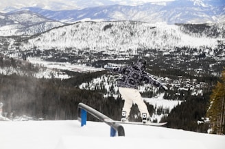 A snowboarder in a patterned winter jacket and white pants is performing a trick on a rail amidst a snowy mountain landscape. The background features expansive, forested mountains covered in snow, under a cloudy sky.
