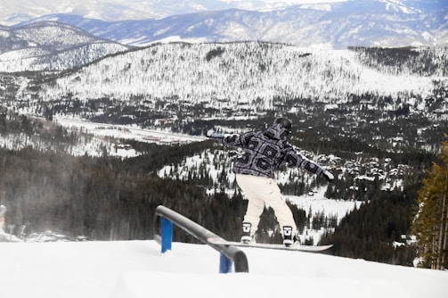 A snowboarder in a patterned winter jacket and white pants is performing a trick on a rail amidst a snowy mountain landscape. The background features expansive, forested mountains covered in snow, under a cloudy sky.