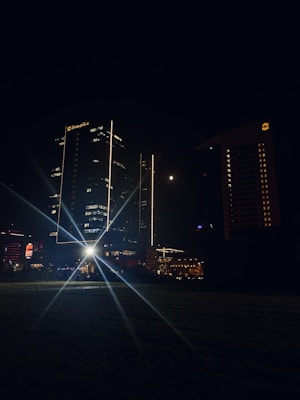A nighttime cityscape showcasing illuminated skyscrapers. The central focus is a tall building with bright vertical lights and a clearly visible hotel logo. A prominent starburst effect emanates from a light source near the ground, adding a dramatic flair. The moon is visible in the sky, adding to the atmosphere.