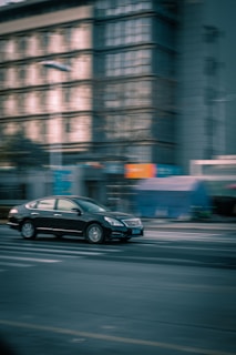 A black car is in motion on an urban street, with the background of a multi-story building showing a sense of speed and dynamism due to the blurring effect.