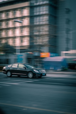 A black car is in motion on an urban street, with the background of a multi-story building showing a sense of speed and dynamism due to the blurring effect.