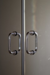 Two parallel stainless steel door handles with black rubber grips mounted on a reflective glass or metallic surface. The handles are positioned symmetrically along a central divider. Subtle lighting creates a soft reflection and a hint of a rainbow effect on the surface.
