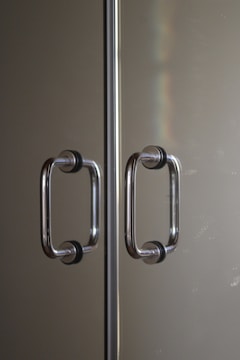 Two parallel stainless steel door handles with black rubber grips mounted on a reflective glass or metallic surface. The handles are positioned symmetrically along a central divider. Subtle lighting creates a soft reflection and a hint of a rainbow effect on the surface.