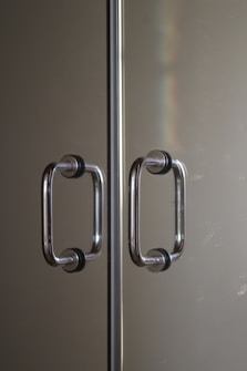 Two parallel stainless steel door handles with black rubber grips mounted on a reflective glass or metallic surface. The handles are positioned symmetrically along a central divider. Subtle lighting creates a soft reflection and a hint of a rainbow effect on the surface.