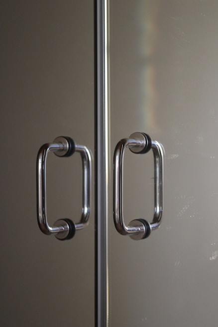 Two parallel stainless steel door handles with black rubber grips mounted on a reflective glass or metallic surface. The handles are positioned symmetrically along a central divider. Subtle lighting creates a soft reflection and a hint of a rainbow effect on the surface.