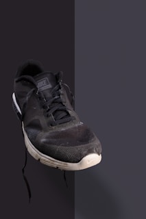 A single black athletic shoe with a white sole is positioned against a plain, dark background. The shoe has visible wear and tear, including scuffs and dirt marks, particularly on the toe area. The laces appear loose, and the shoe is slightly angled to show its front and side.