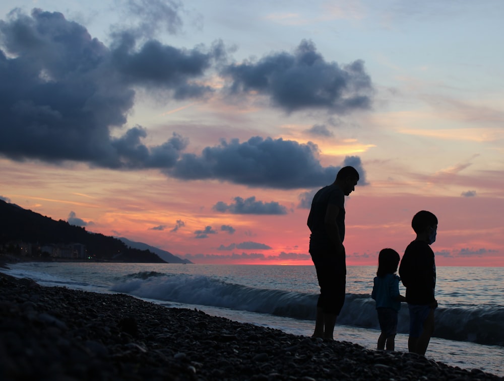 silhouette of 2 men and woman standing on seashore during sunset