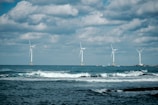 Four wind turbines stand across the open sea with waves crashing in the foreground. The sky is partially cloudy, creating a dramatic background for the clean energy structures.