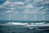 Four wind turbines stand across the open sea with waves crashing in the foreground. The sky is partially cloudy, creating a dramatic background for the clean energy structures.