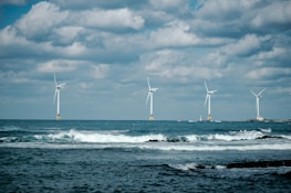 Four wind turbines stand across the open sea with waves crashing in the foreground. The sky is partially cloudy, creating a dramatic background for the clean energy structures.