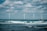 Four wind turbines stand across the open sea with waves crashing in the foreground. The sky is partially cloudy, creating a dramatic background for the clean energy structures.