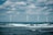 Four wind turbines stand across the open sea with waves crashing in the foreground. The sky is partially cloudy, creating a dramatic background for the clean energy structures.