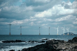 Several offshore wind turbines are positioned in a line across the sea, beneath a cloudy sky. The foreground features rocky coastline with waves crashing against the shore.