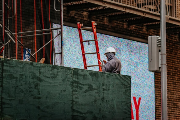 A construction worker wearing a hard hat and face mask is standing near a red ladder and scaffolding, appearing to use a mobile device. The background includes a digital screen displaying static noise, with some brick wall visible.