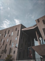 A modern building with a geometric design featuring large, vertical windows and an angled roof structure. The facade is a combination of concrete and glass elements, reflecting a cloudy sky above. A billboard with a fashion advertisement is visible, adding a touch of color and contrast.