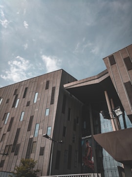 A modern building with a geometric design featuring large, vertical windows and an angled roof structure. The facade is a combination of concrete and glass elements, reflecting a cloudy sky above. A billboard with a fashion advertisement is visible, adding a touch of color and contrast.