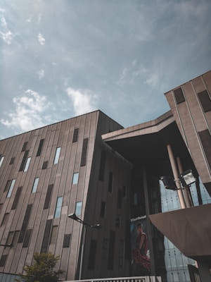 A modern building with a geometric design featuring large, vertical windows and an angled roof structure. The facade is a combination of concrete and glass elements, reflecting a cloudy sky above. A billboard with a fashion advertisement is visible, adding a touch of color and contrast.