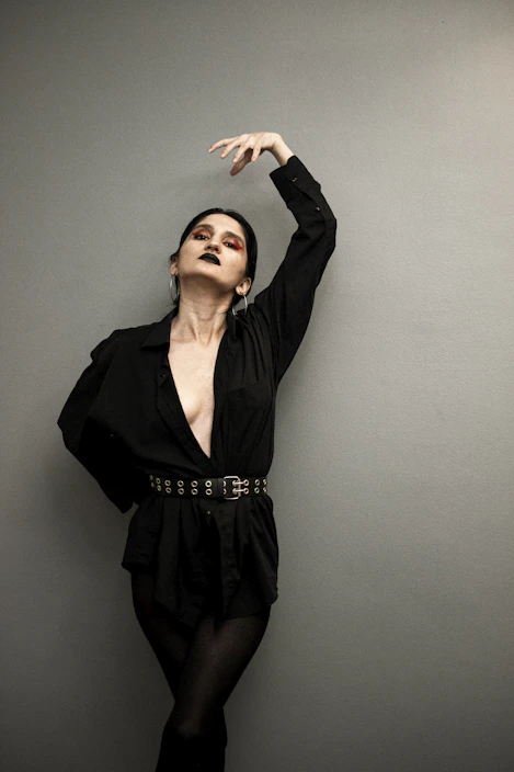 A dark, moody photo of a model wearing gothic clothing with intricate leather and lace details, standing against a textured black wall.