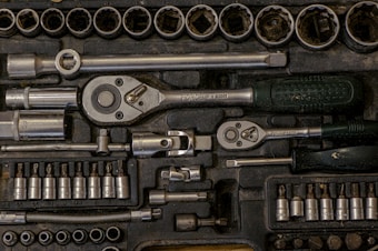 A collection of various metal tools neatly organized in a black foam case. Included are ratchets, sockets, and other attachments, each in their designated slot. The tools appear to be primarily made of metal, with some having rubberized handles.