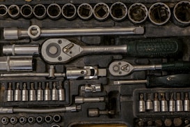 A collection of various metal tools neatly organized in a black foam case. Included are ratchets, sockets, and other attachments, each in their designated slot. The tools appear to be primarily made of metal, with some having rubberized handles.