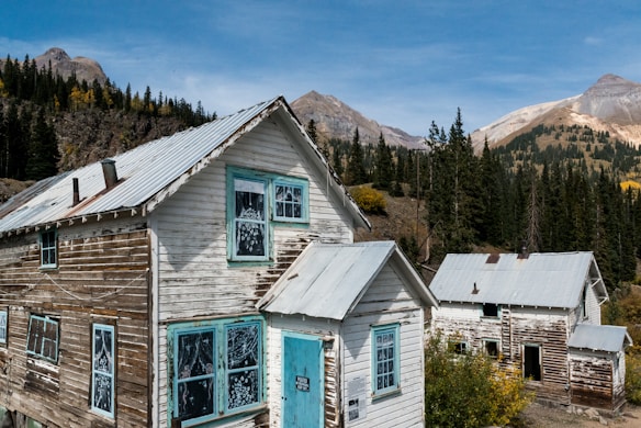 Two rustic wooden houses with white paint and blue trim are situated in a mountainous and forested landscape. The buildings have tin roofs and display signs of weathering and age, with peeling paint and visible wood. In the background, there are tall evergreen trees and distant mountains under a clear blue sky.