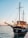 A large, elegant sailing boat is docked by the pier at sunset. The boat features tall masts, a sleek black hull, and a wooden deck with railings. A small flag is seen waving at the front, with large fenders hanging over the side. The sky is painted with soft hues of pink and blue, reflecting on the tranquil water.