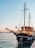A large, elegant sailing boat is docked by the pier at sunset. The boat features tall masts, a sleek black hull, and a wooden deck with railings. A small flag is seen waving at the front, with large fenders hanging over the side. The sky is painted with soft hues of pink and blue, reflecting on the tranquil water.