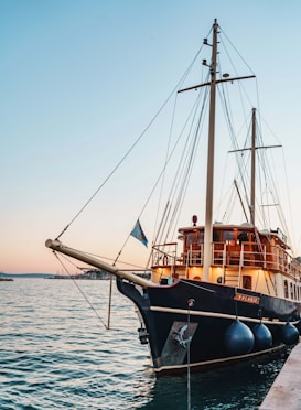 A large, elegant sailing boat is docked by the pier at sunset. The boat features tall masts, a sleek black hull, and a wooden deck with railings. A small flag is seen waving at the front, with large fenders hanging over the side. The sky is painted with soft hues of pink and blue, reflecting on the tranquil water.