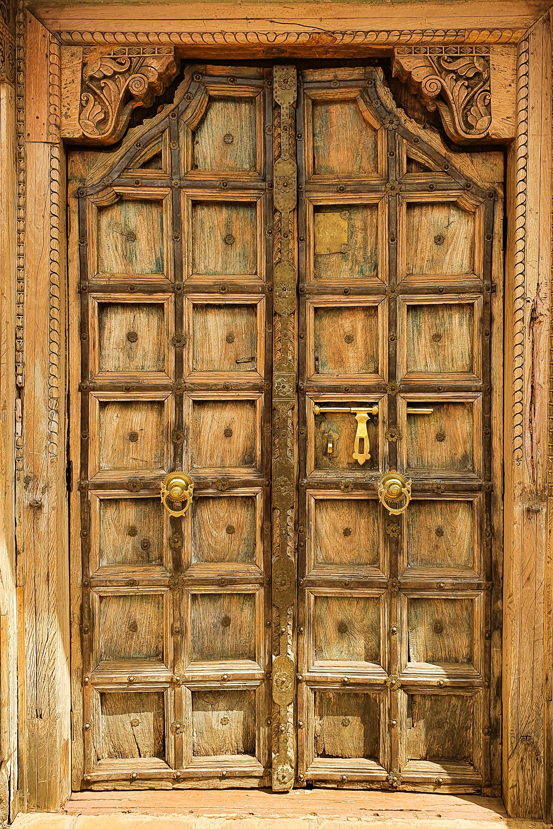 An intricately carved wooden door featuring ornate details and a rustic appearance. It has a grid pattern with small square panels, metal studs, and decorative metal handles. The frame is embellished with carved floral designs, conveying an antique and artistic style.