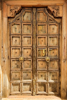 An intricately carved wooden door featuring ornate details and a rustic appearance. It has a grid pattern with small square panels, metal studs, and decorative metal handles. The frame is embellished with carved floral designs, conveying an antique and artistic style.