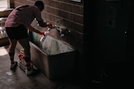 A person wearing a red and white striped soccer uniform fills up a white bottle from a faucet above an old metal trough. The setting appears to be a dimly lit locker room or utility area with concrete floors and brick walls.
