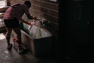 A person wearing a red and white striped soccer uniform fills up a white bottle from a faucet above an old metal trough. The setting appears to be a dimly lit locker room or utility area with concrete floors and brick walls.