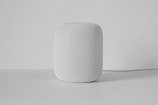 A white cylindrical smart speaker with a mesh exterior sits on a smooth, light-colored surface. The background is plain and minimalistic, emphasizing the device.