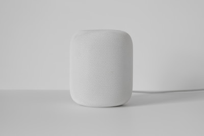 A white cylindrical smart speaker with a mesh exterior sits on a smooth, light-colored surface. The background is plain and minimalistic, emphasizing the device.