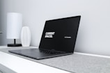 A sleek, modern laptop with a black screen displaying the text 'CURRENT SPACES.' on the left and 'Hi, I'm brandon.' on the right. The laptop is placed on a light gray surface with a minimalist design. A white, textured vase and a white, cylindrical lamp are positioned in the background on a white desk against a light gray wall.