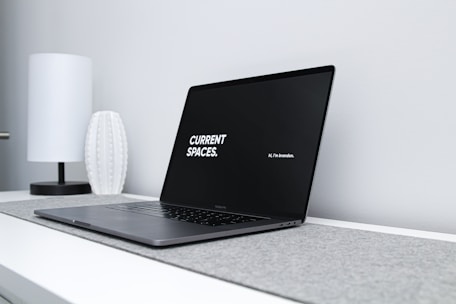 A sleek, modern laptop with a black screen displaying the text 'CURRENT SPACES.' on the left and 'Hi, I'm brandon.' on the right. The laptop is placed on a light gray surface with a minimalist design. A white, textured vase and a white, cylindrical lamp are positioned in the background on a white desk against a light gray wall.