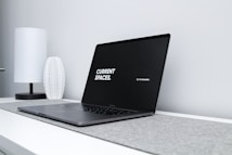 A sleek, modern laptop with a black screen displaying the text 'CURRENT SPACES.' on the left and 'Hi, I'm brandon.' on the right. The laptop is placed on a light gray surface with a minimalist design. A white, textured vase and a white, cylindrical lamp are positioned in the background on a white desk against a light gray wall.