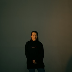 A person stands in a dark room with minimal lighting, wearing a black sweatshirt with white text. The background is a plain, dark green wall creating a mysterious and somber atmosphere. The person has minimal facial expressions and appears to be posing calmly with arms crossed at the waist.